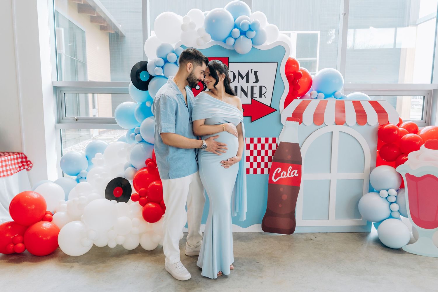 A couple embraces at a baby shower decorated with red and blue balloons and a retro diner theme backdrop.