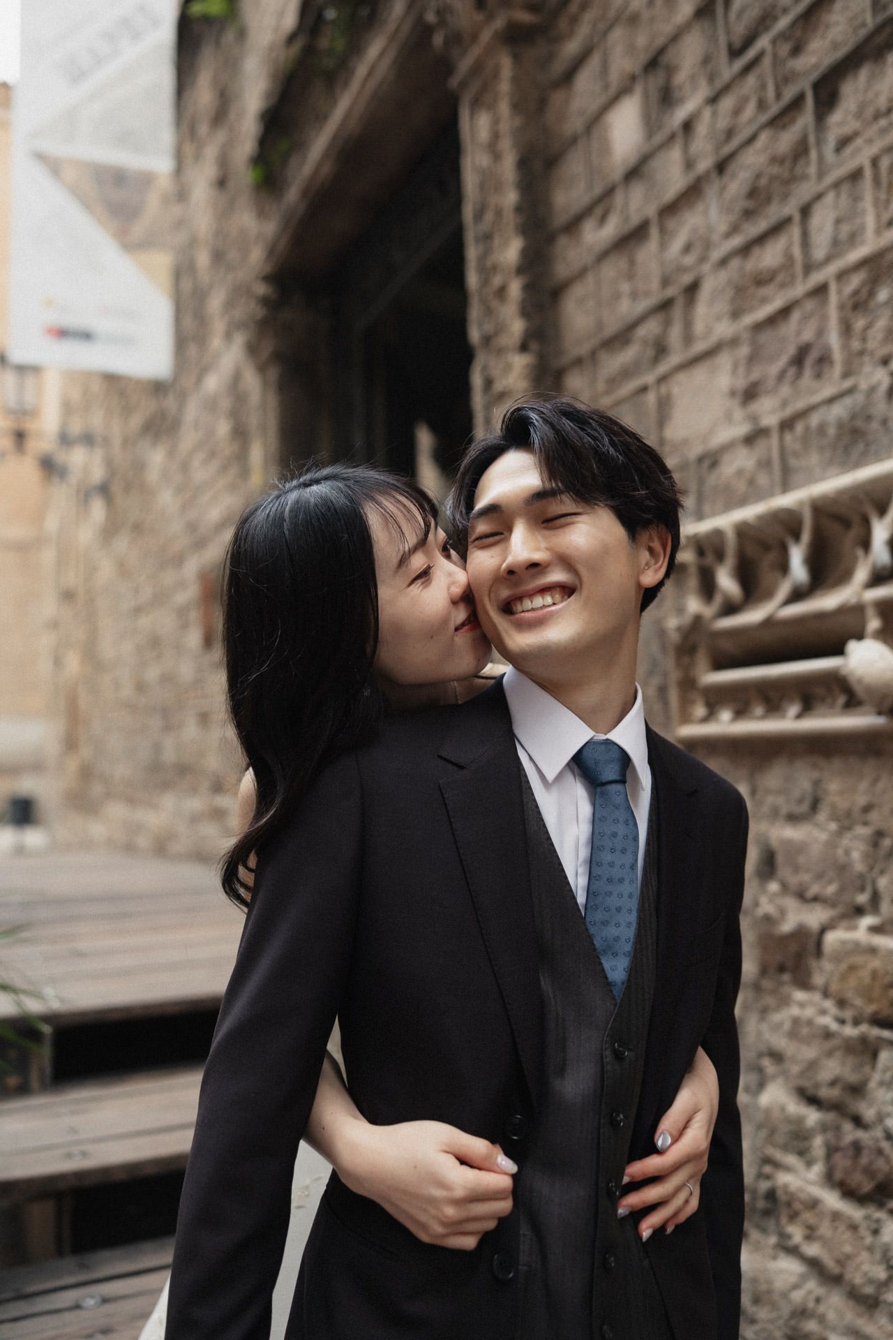 A couple in formal attire share an intimate moment against a stone wall backdrop.