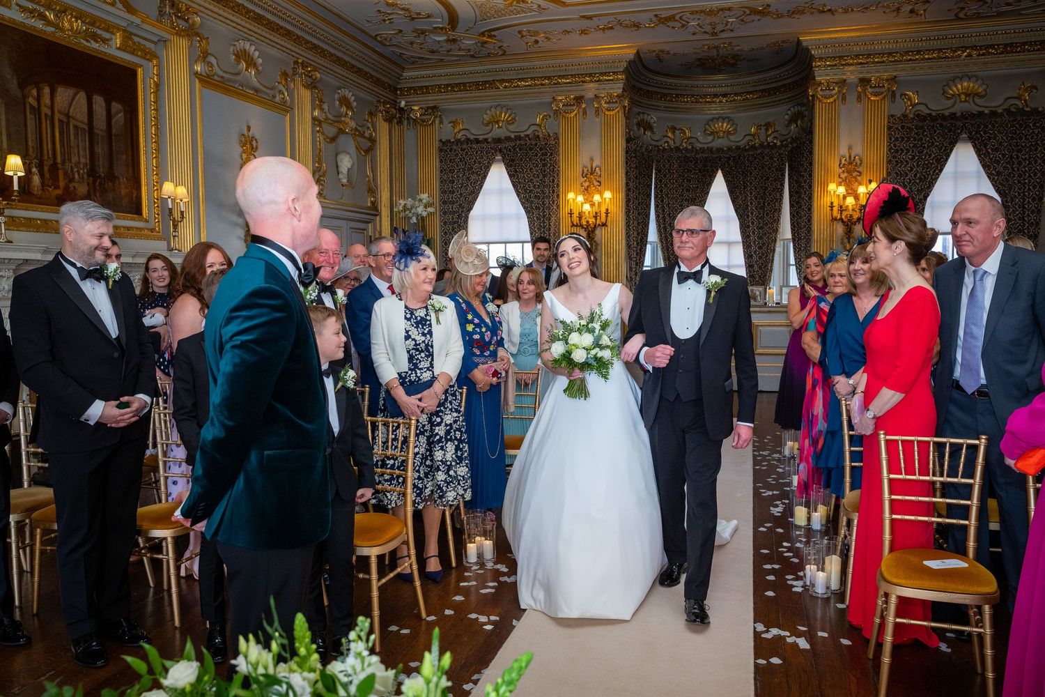 Bride walking down the aisle with her father toward the groom in an elegant wedding ceremony room.