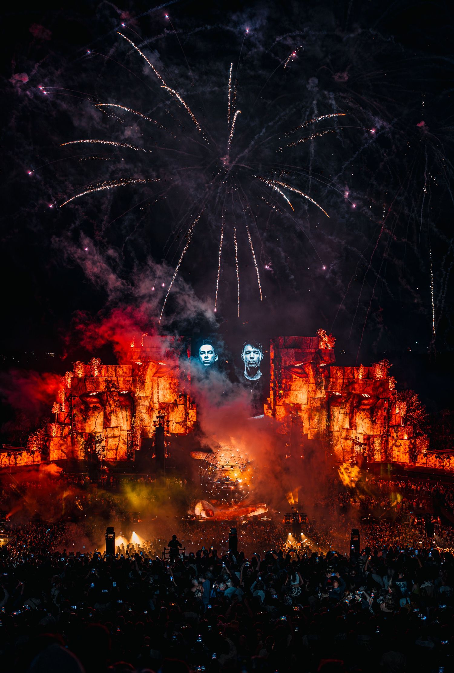 Massive festival stage with fireworks, fire effects, skull projections, and large crowd at night.