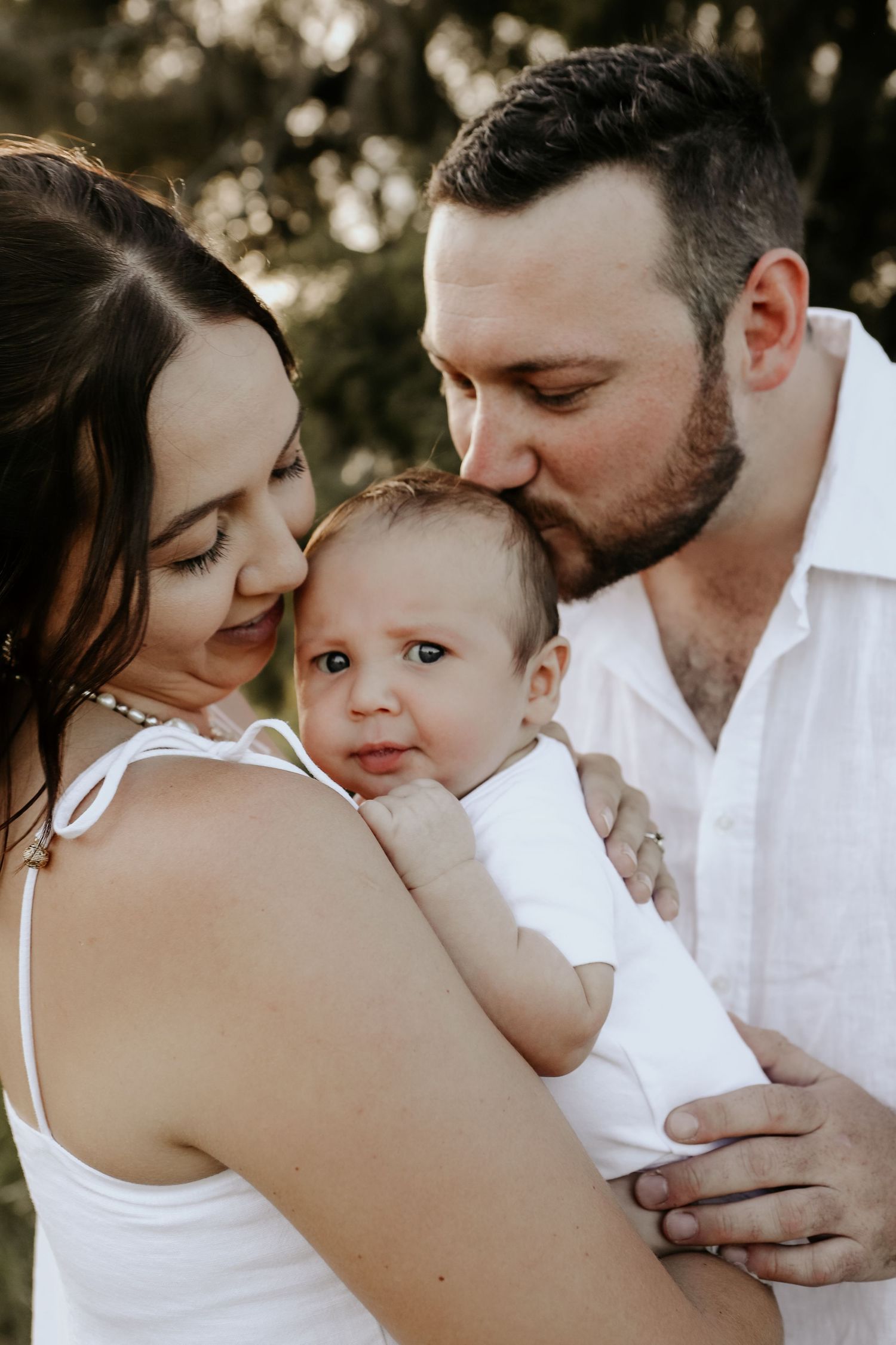 A tender family moment captured outdoors as parents share an intimate moment with their baby in white clothing.