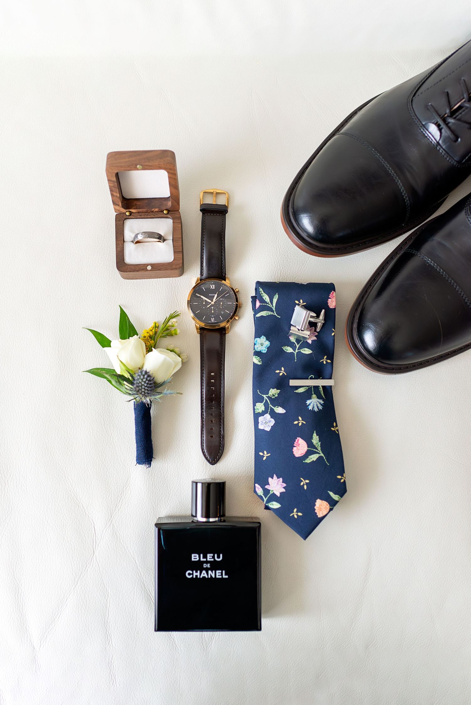 Groom wedding accessories flatlay featuring Bleu de Chanel cologne, floral tie, watch, boutonniere, ring box, and black dress shoes.