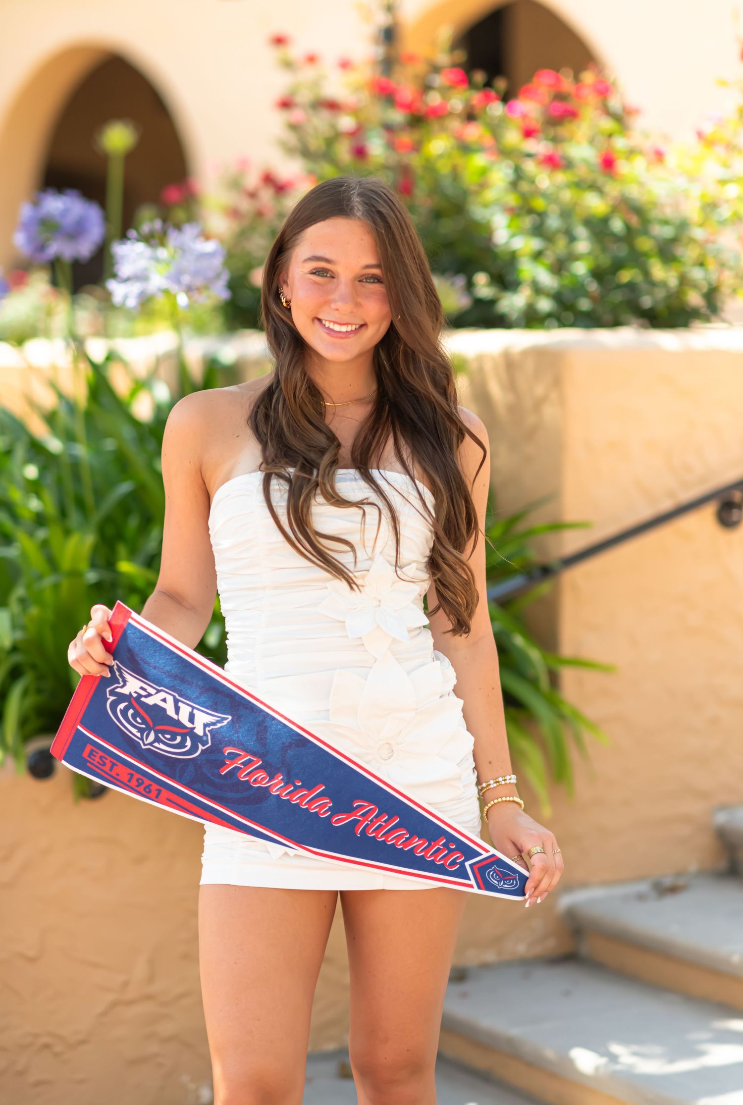 Young woman in white dress smiling and holding a Florida Atlantic University FAU pennant outdoors.