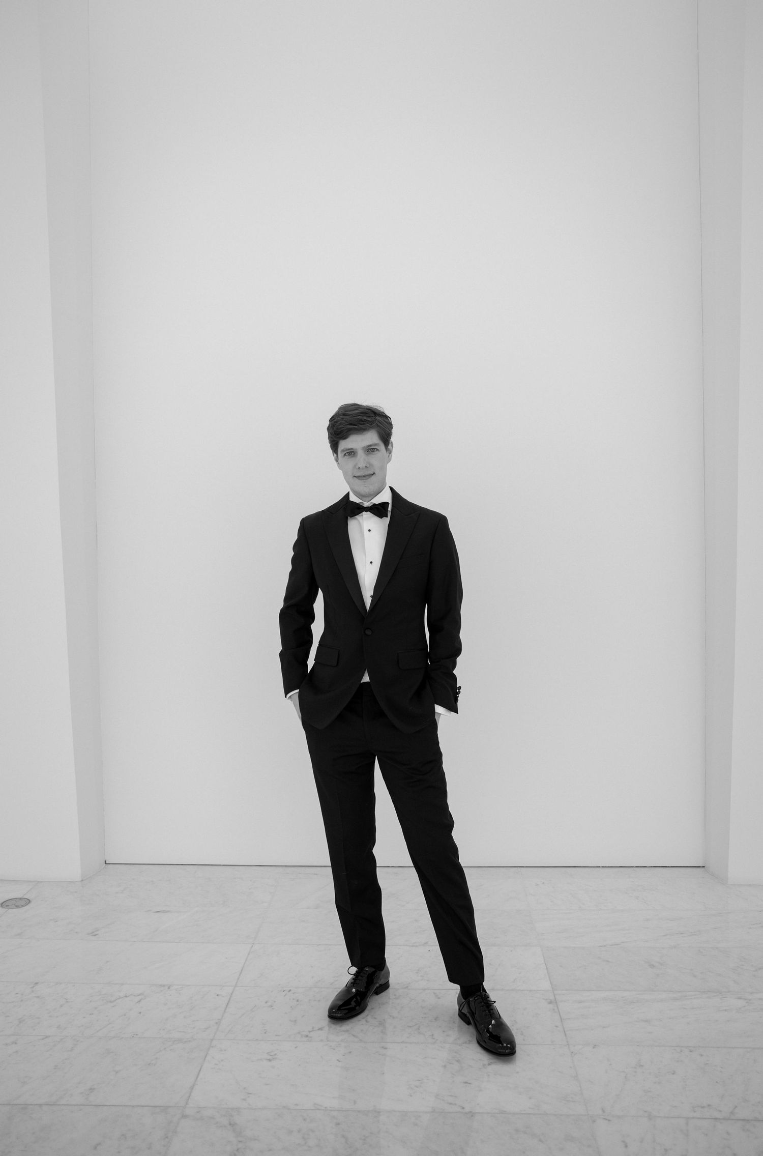Young man in black tuxedo with bow tie standing confidently against white wall in black and white photo.