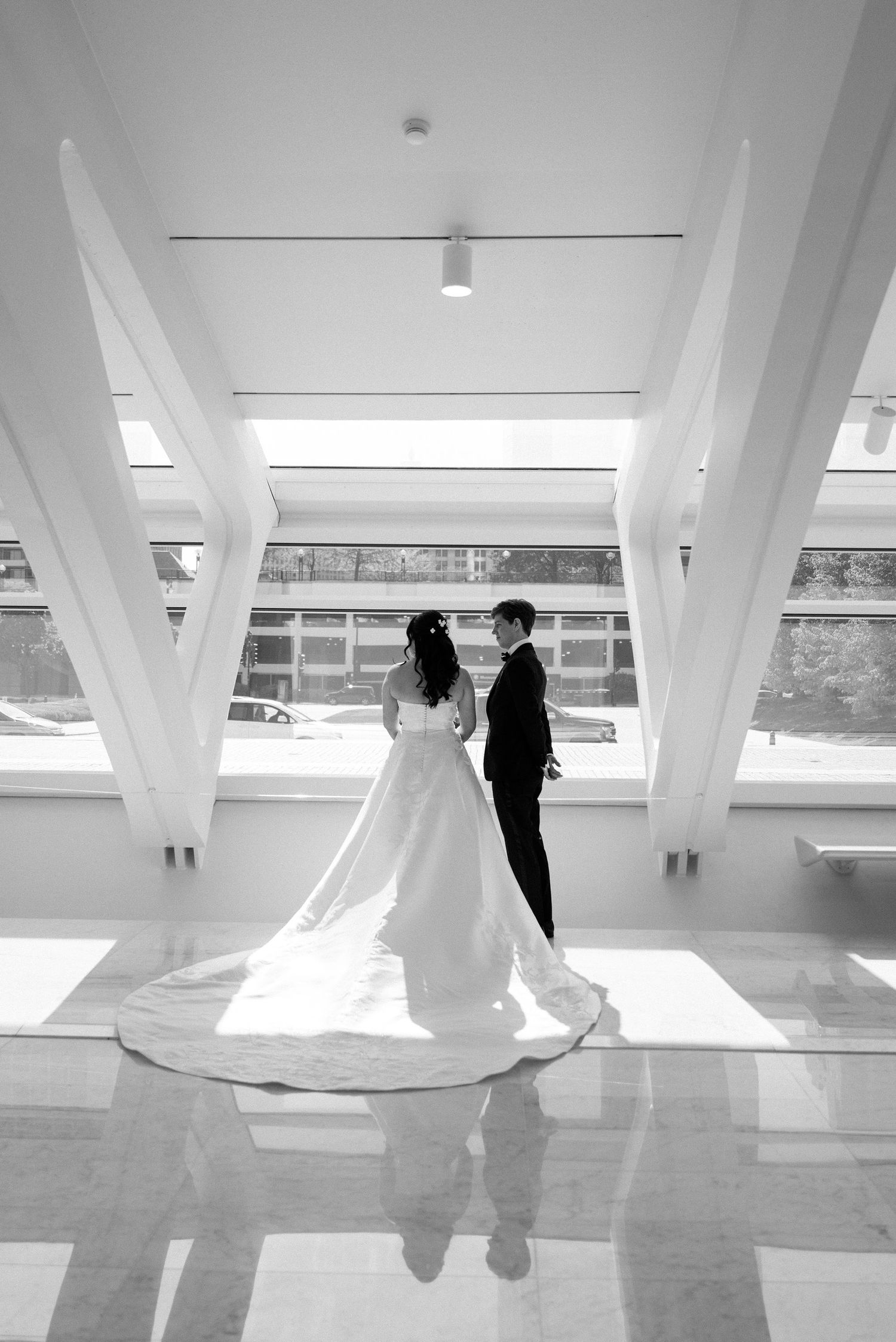 Black and white photo of bride and groom standing together in elegant sunlit hall with marble floors.