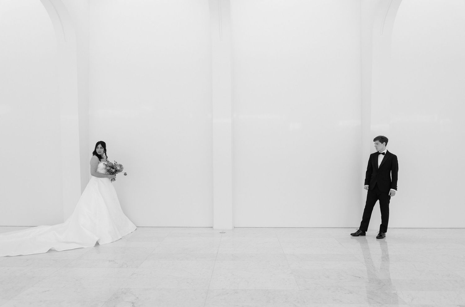 Black and white wedding photo of bride in white gown and groom in tuxedo standing apart in minimalist white gallery space.