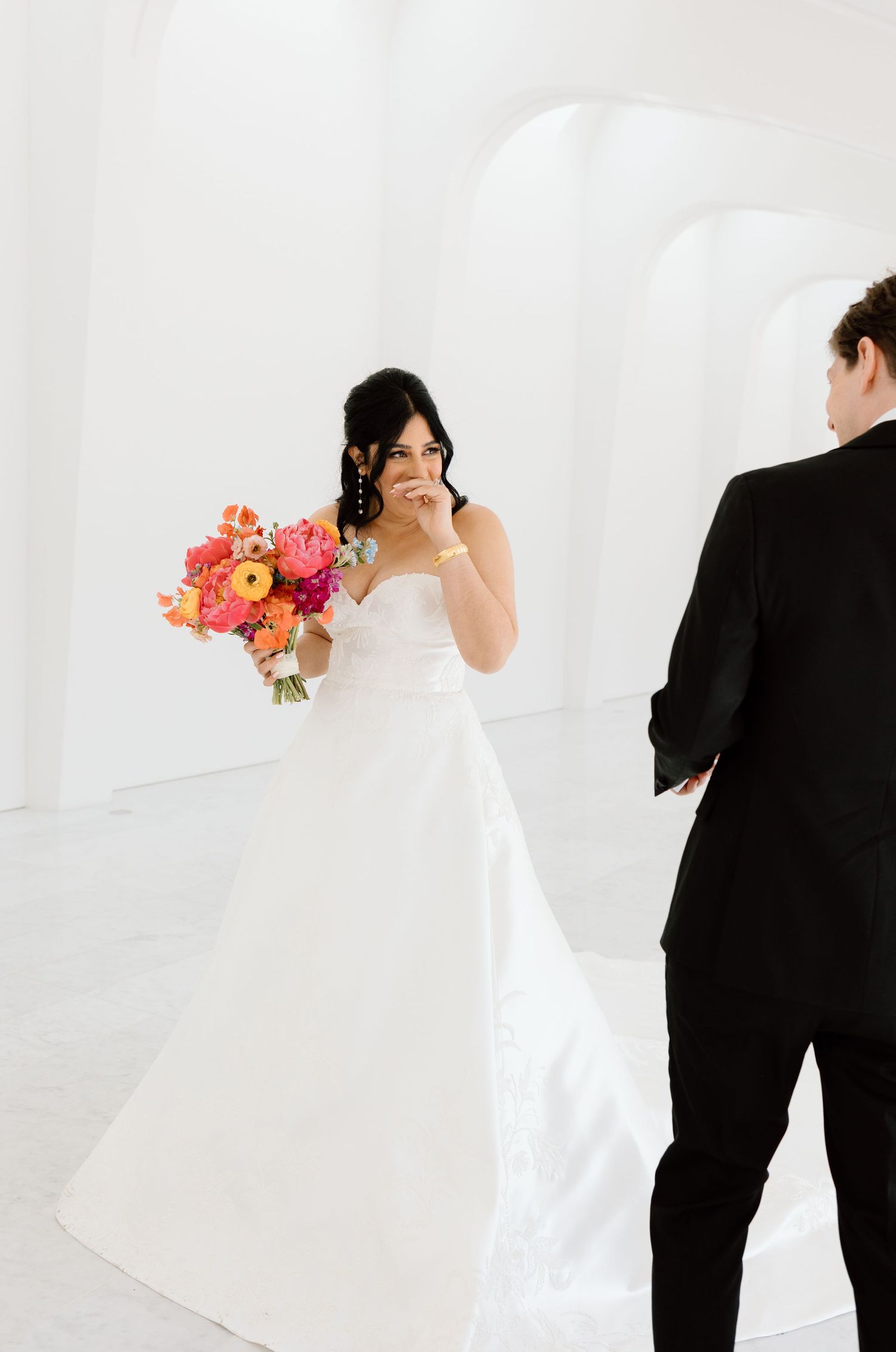 Bride in strapless white gown holds vibrant pink and orange bouquet during emotional first look with groom in black tuxedo.