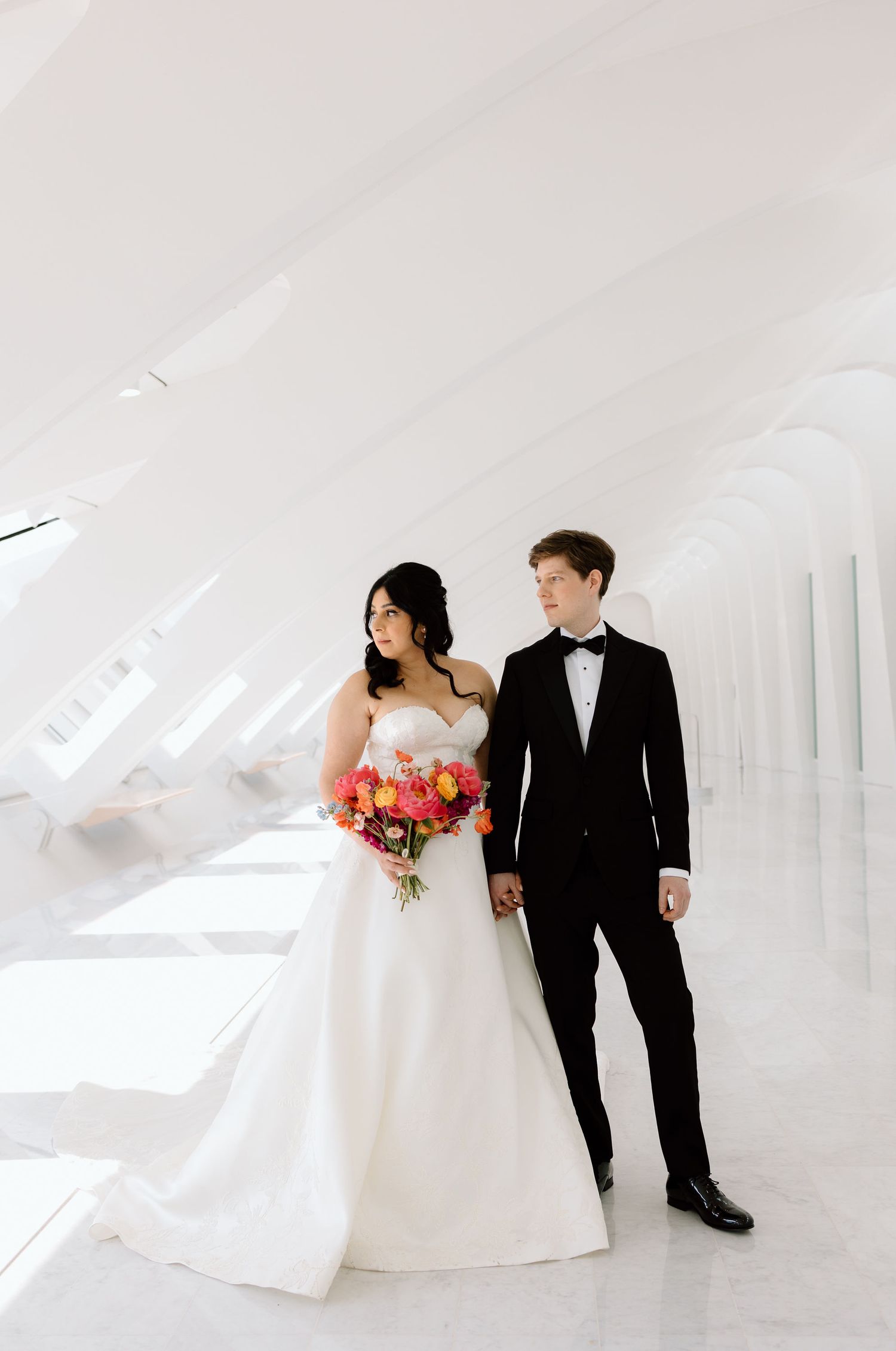 Bride in white gown holding colorful bouquet stands with groom in black tuxedo in a bright modern white venue.
