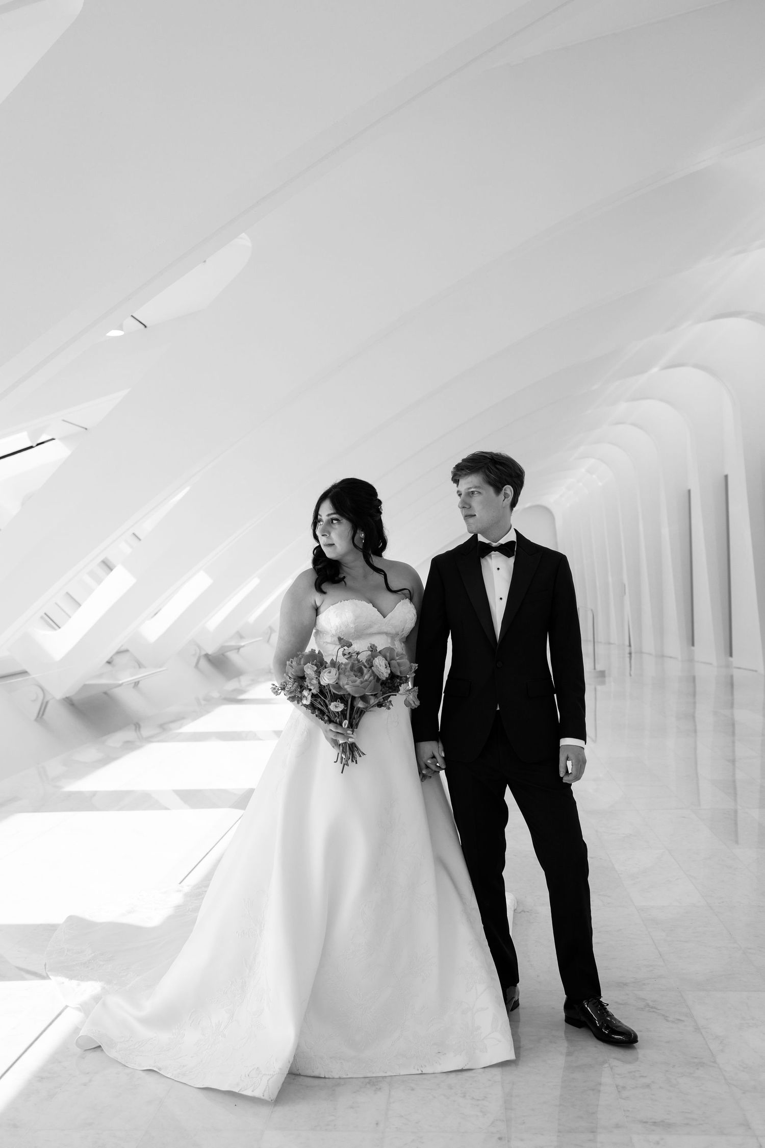 Bride and groom pose elegantly in black and white photo inside a modern architectural wedding venue with arched columns.