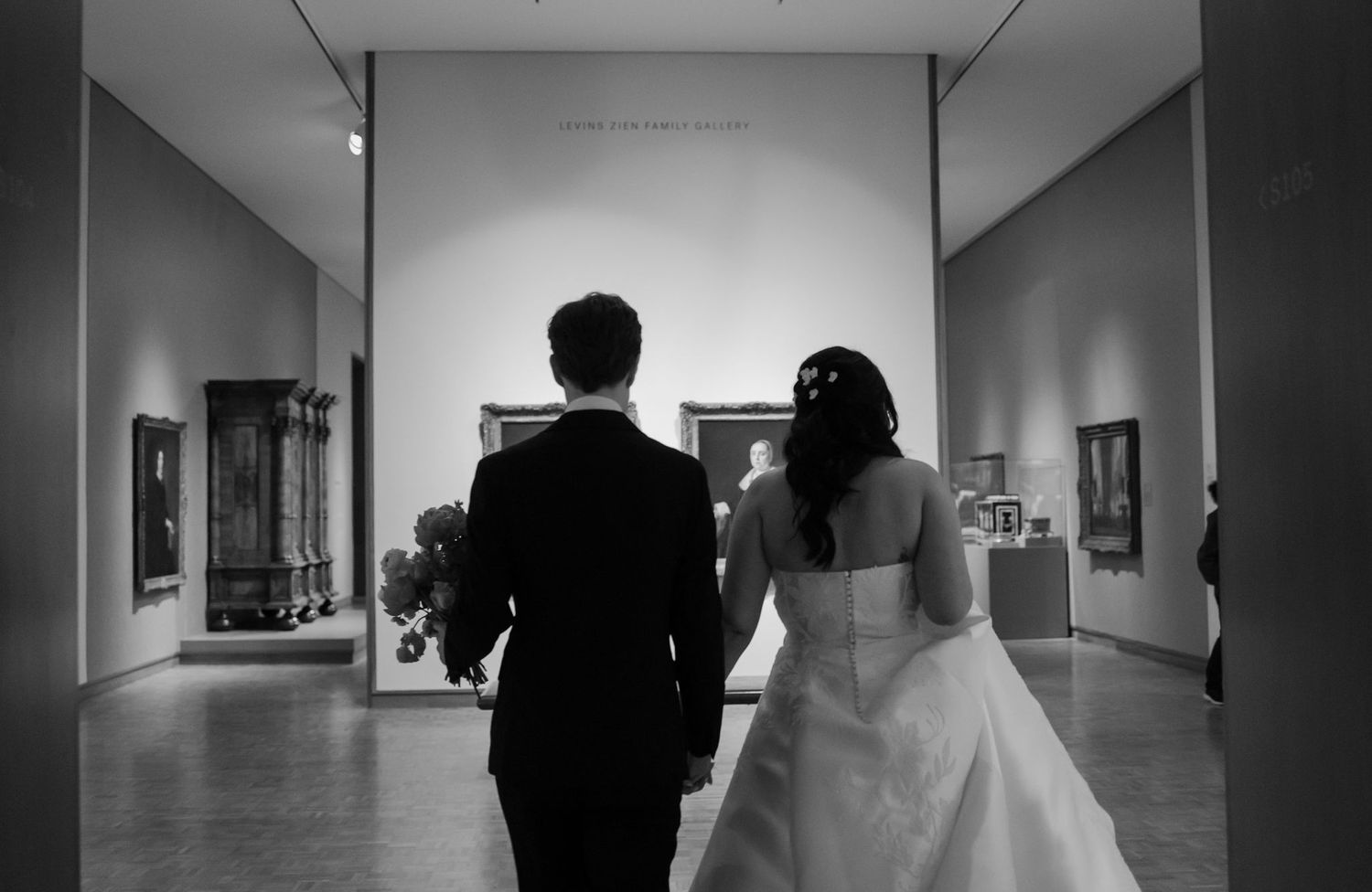 Black and white photo of a bride and groom walking hand in hand through an art gallery on their wedding day.
