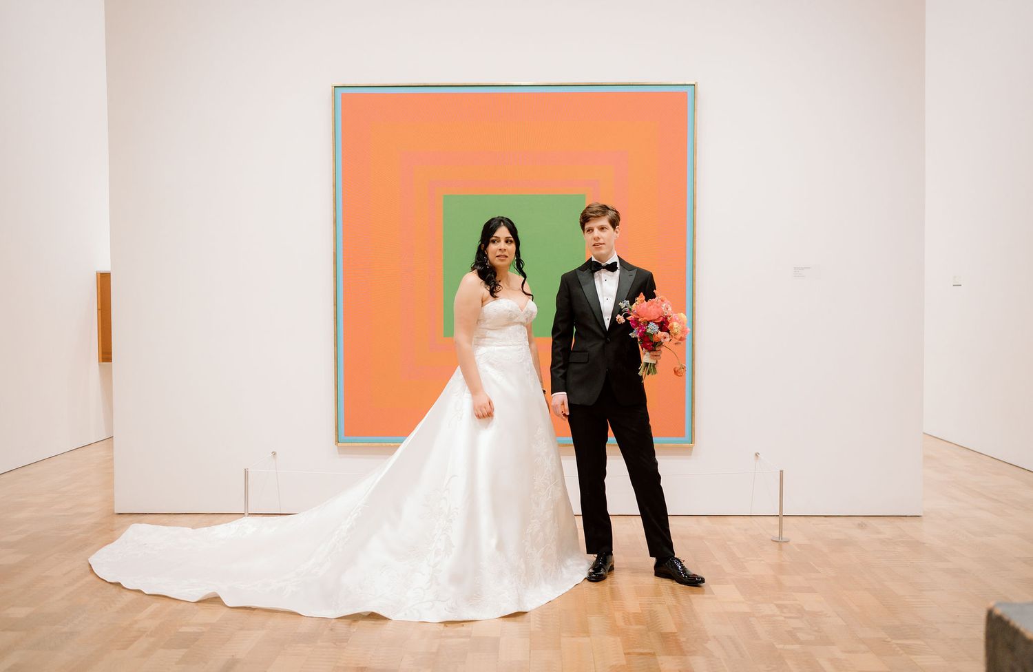 Bride and groom posing in white gown and tuxedo before a large colorful abstract painting in a modern art museum.