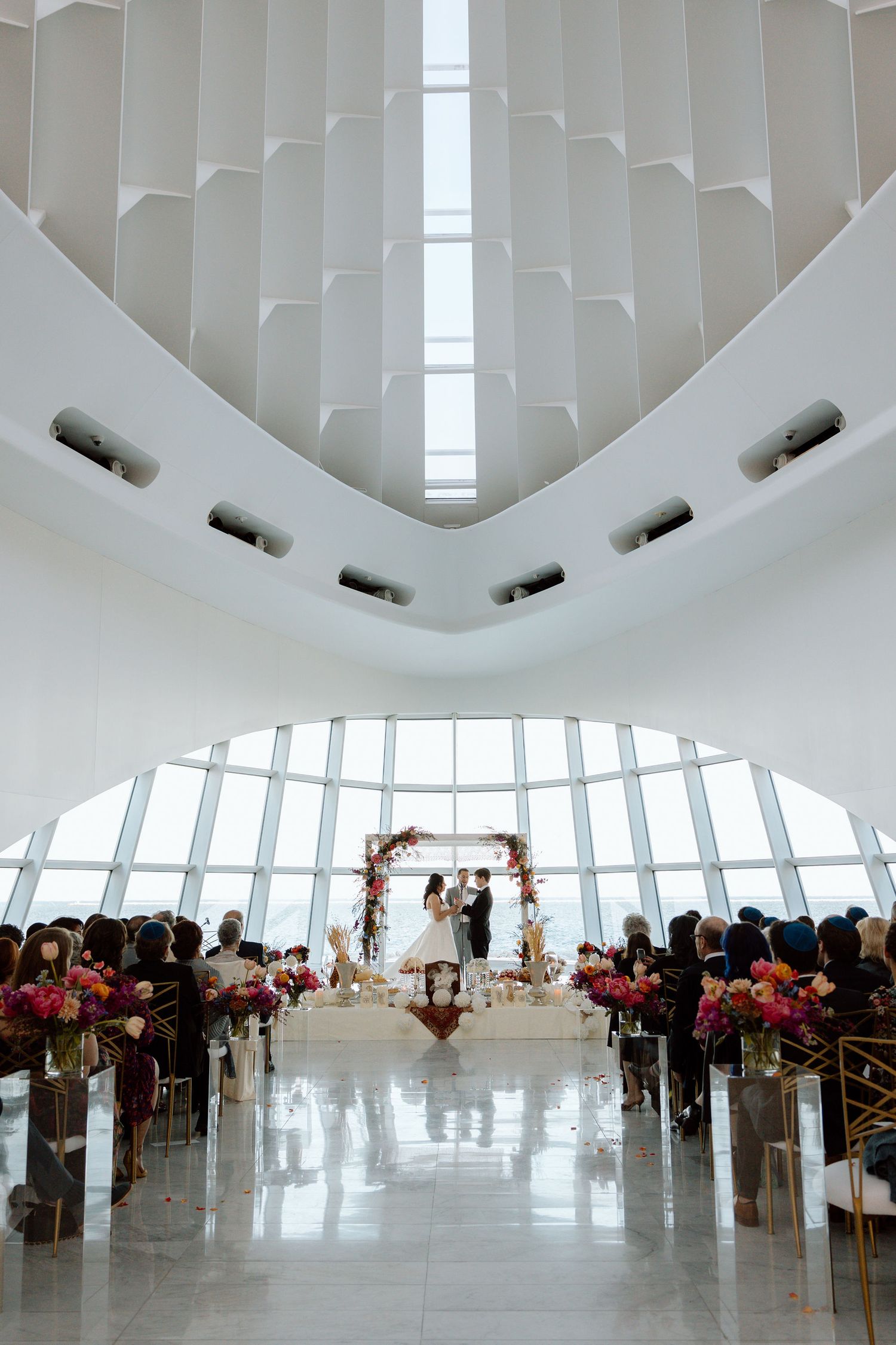 Elegant wedding ceremony in a modern white architectural venue with floral arch, colorful blooms lining the aisle.