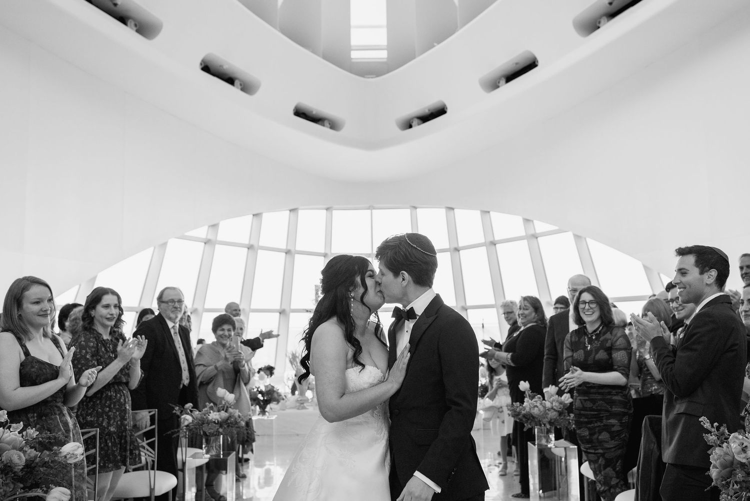Bride and groom share first kiss at altar surrounded by applauding guests in a stunning modern museum wedding venue.