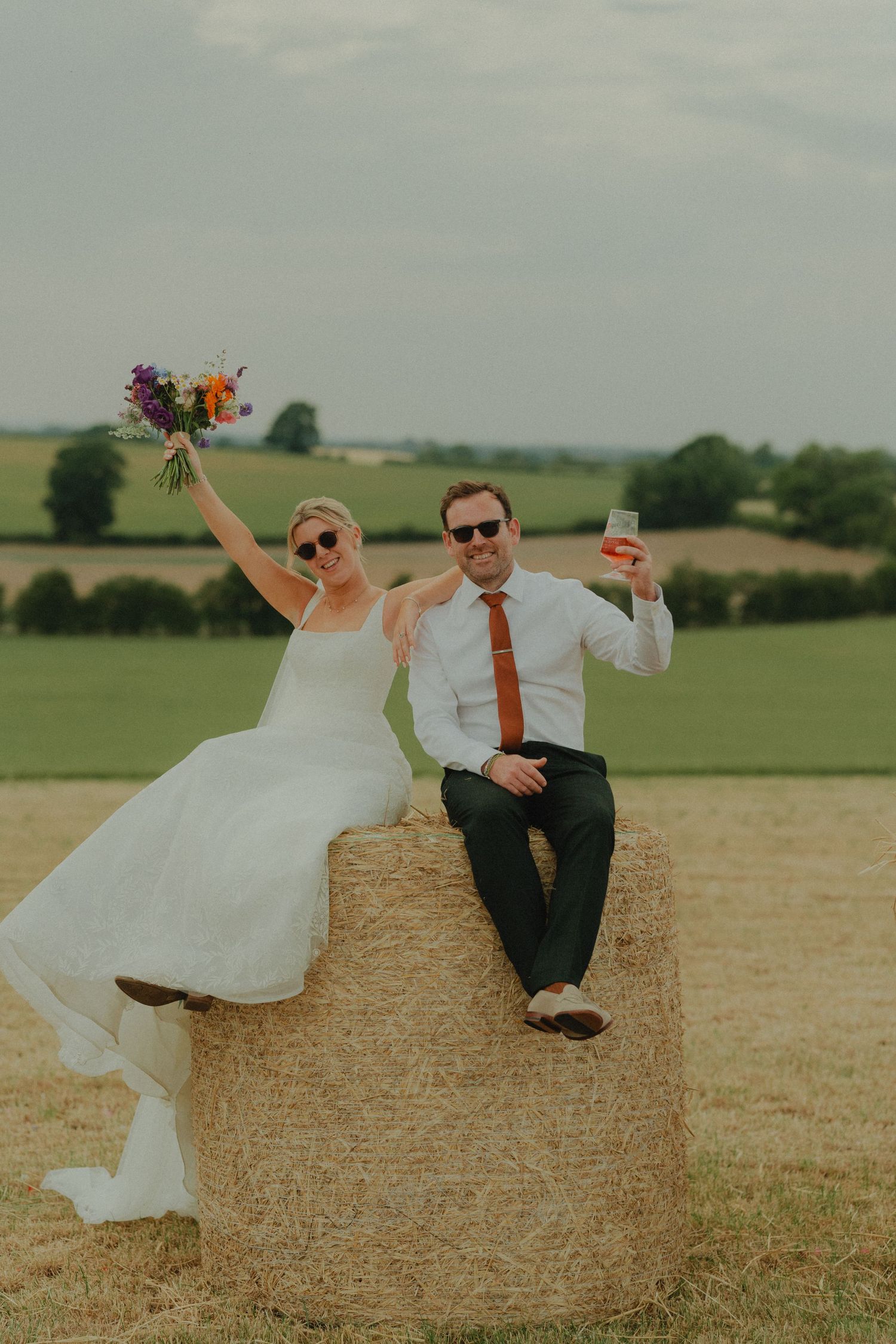 Bride and groom sitting on a hay bale in a field, raising bouquet and drink, wearing sunglasses on their wedding day.
