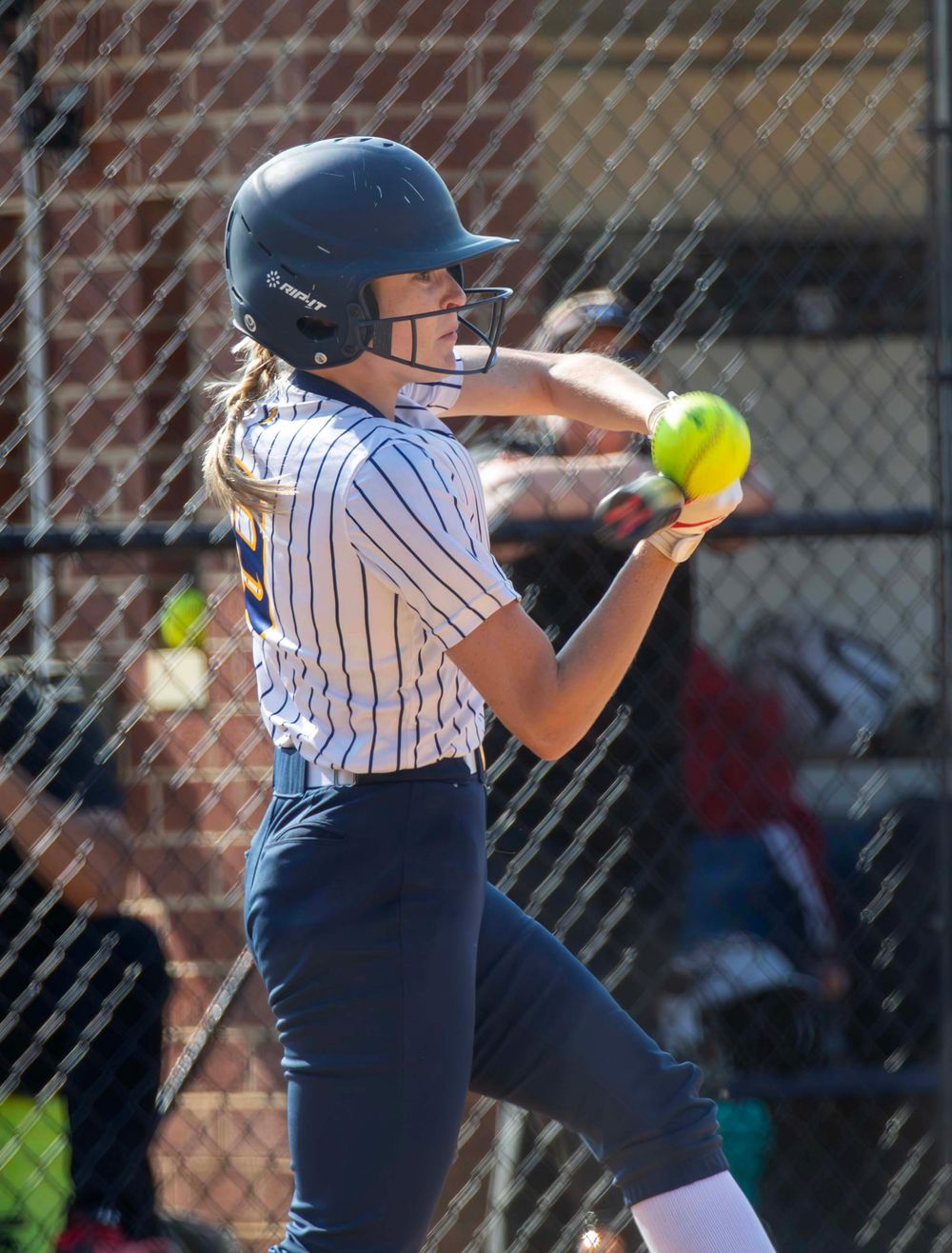 Softball player in pinstriped uniform swings bat at yellow ball during game action.