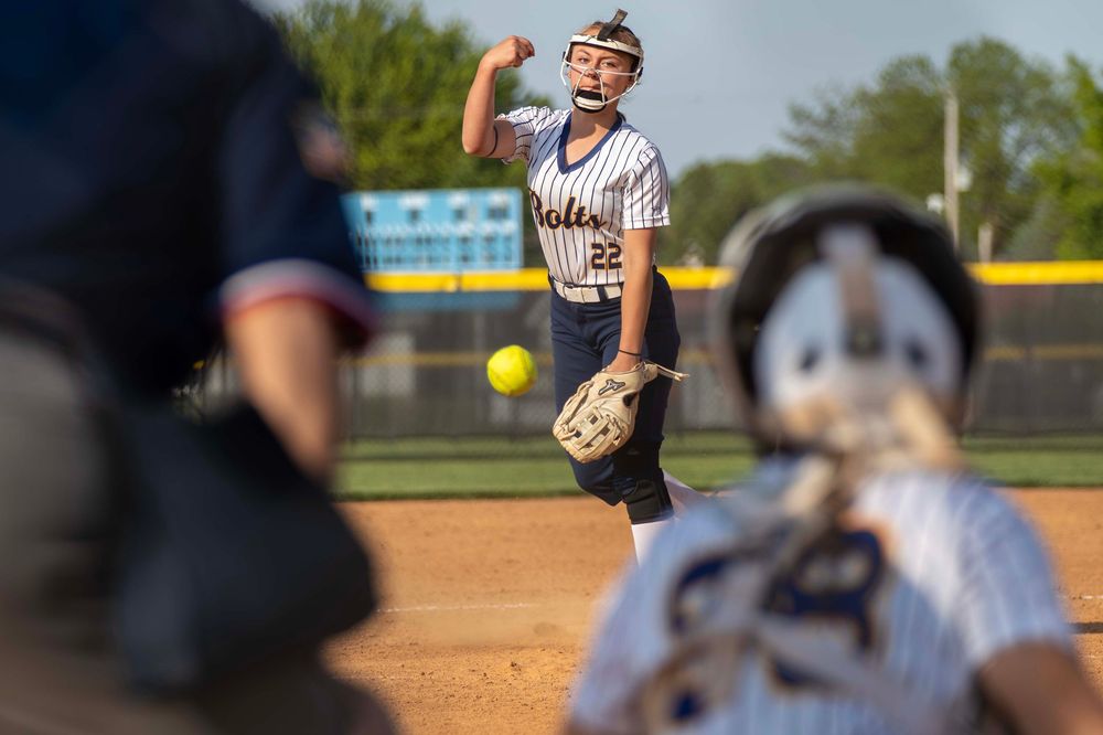 A softball pitcher releases the ball during a game with a focused expression and determined stance on the field.