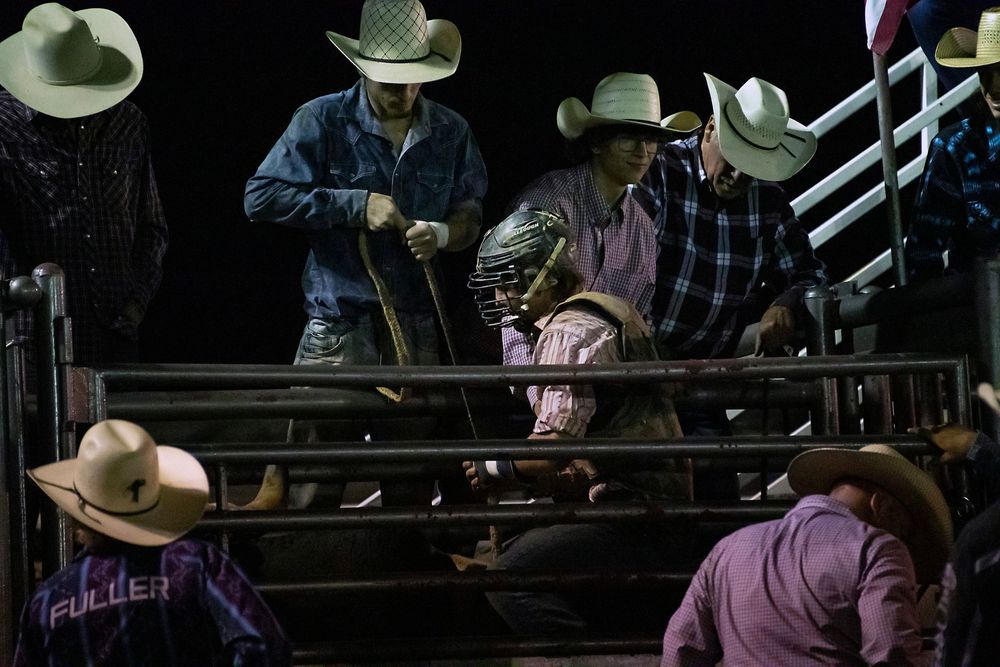 Rodeo cowboys in cowboy hats watch intently from behind metal gates at a nighttime arena event.