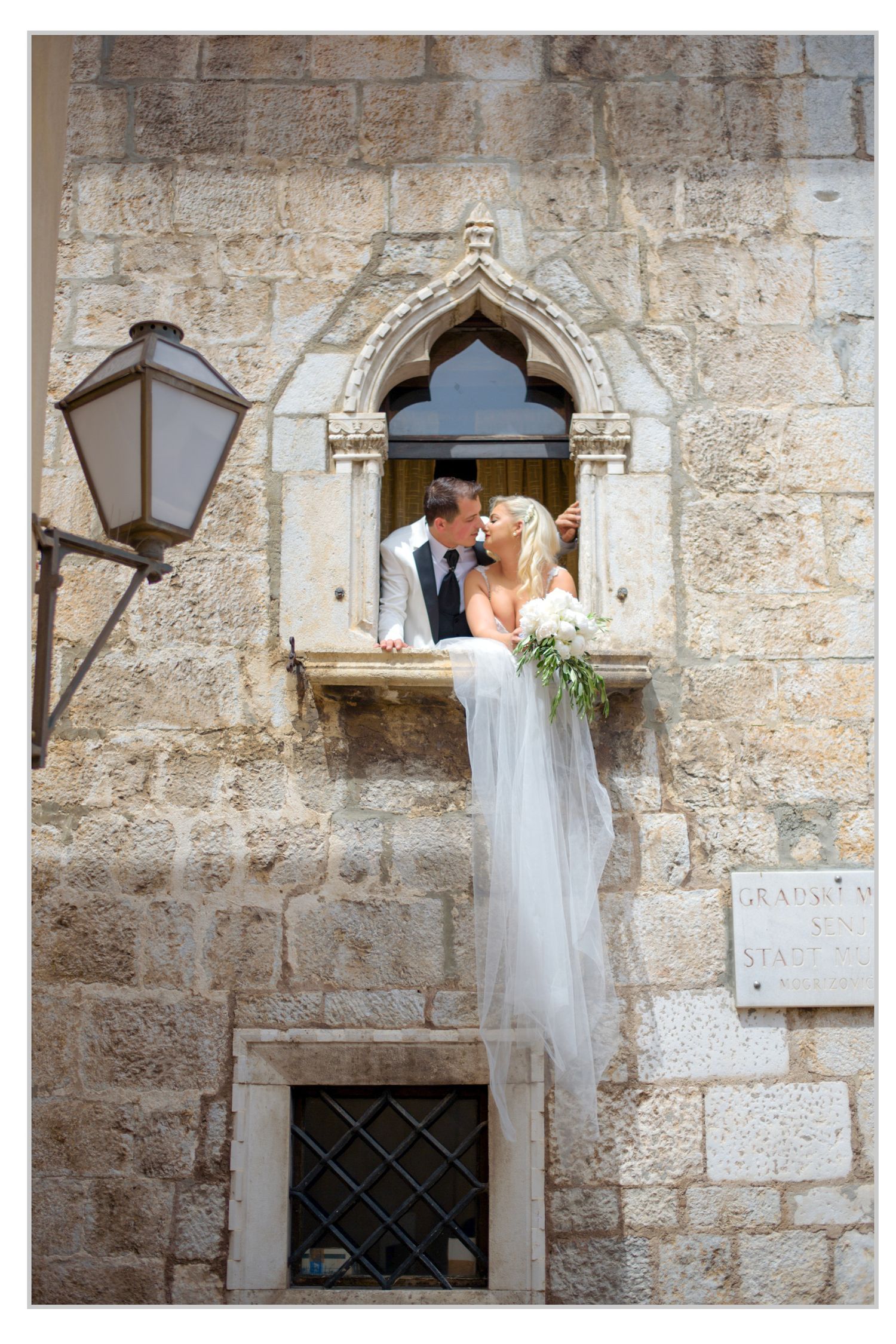 A romantic wedding moment captured through a stone archway window with a decorative lantern against an ancient wall.