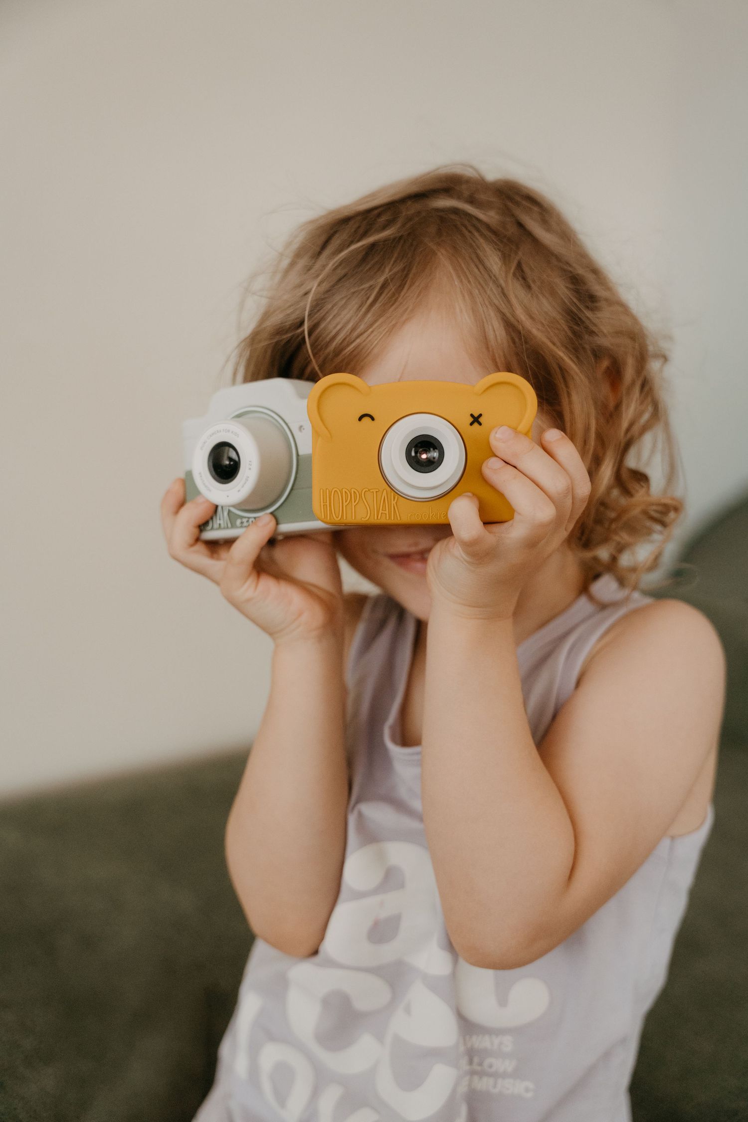 A young girl with curly hair is holding a yellow toy camera and looking through the viewfinder.