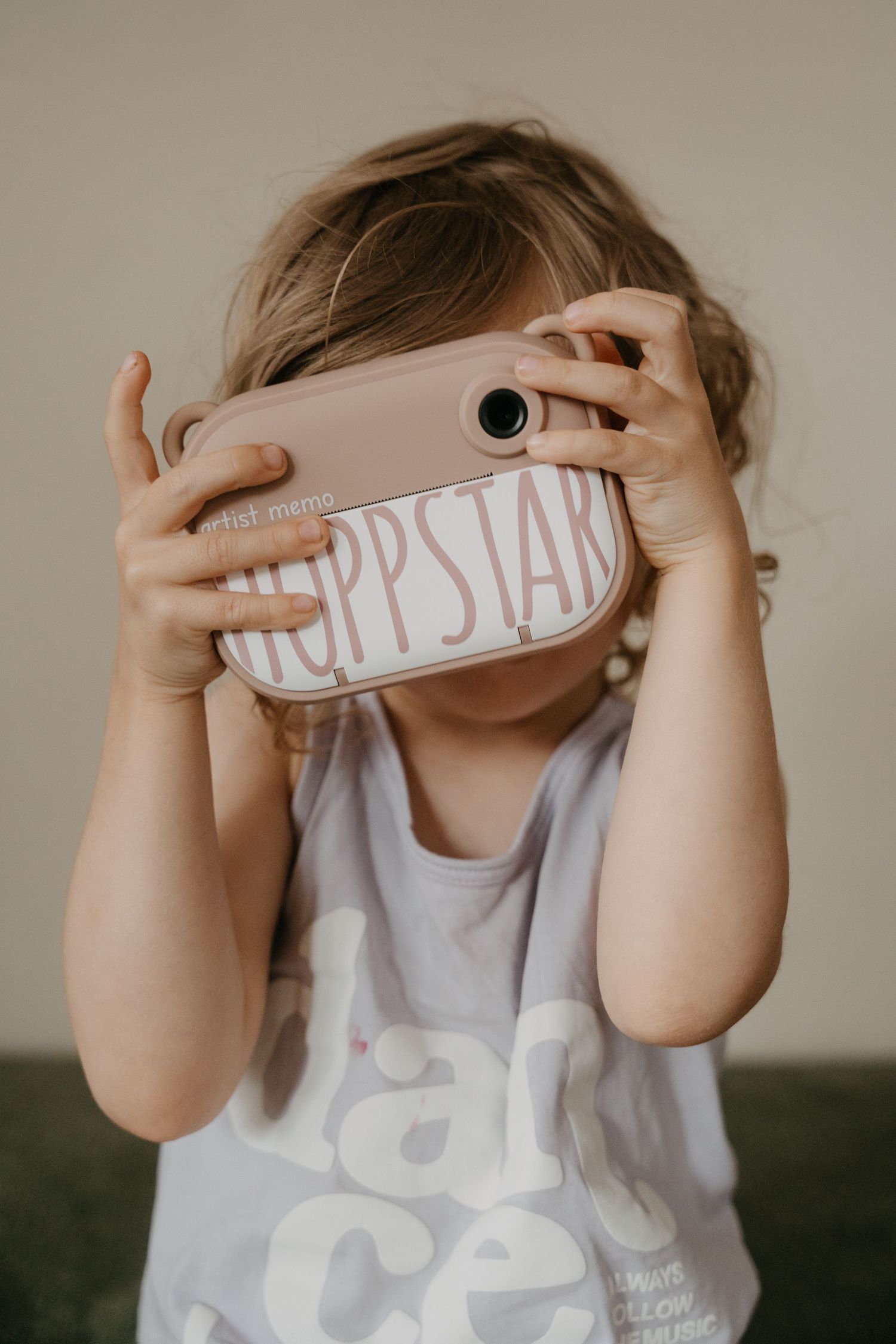 A young girl with curly hair is holding a yellow toy camera and looking through the viewfinder.