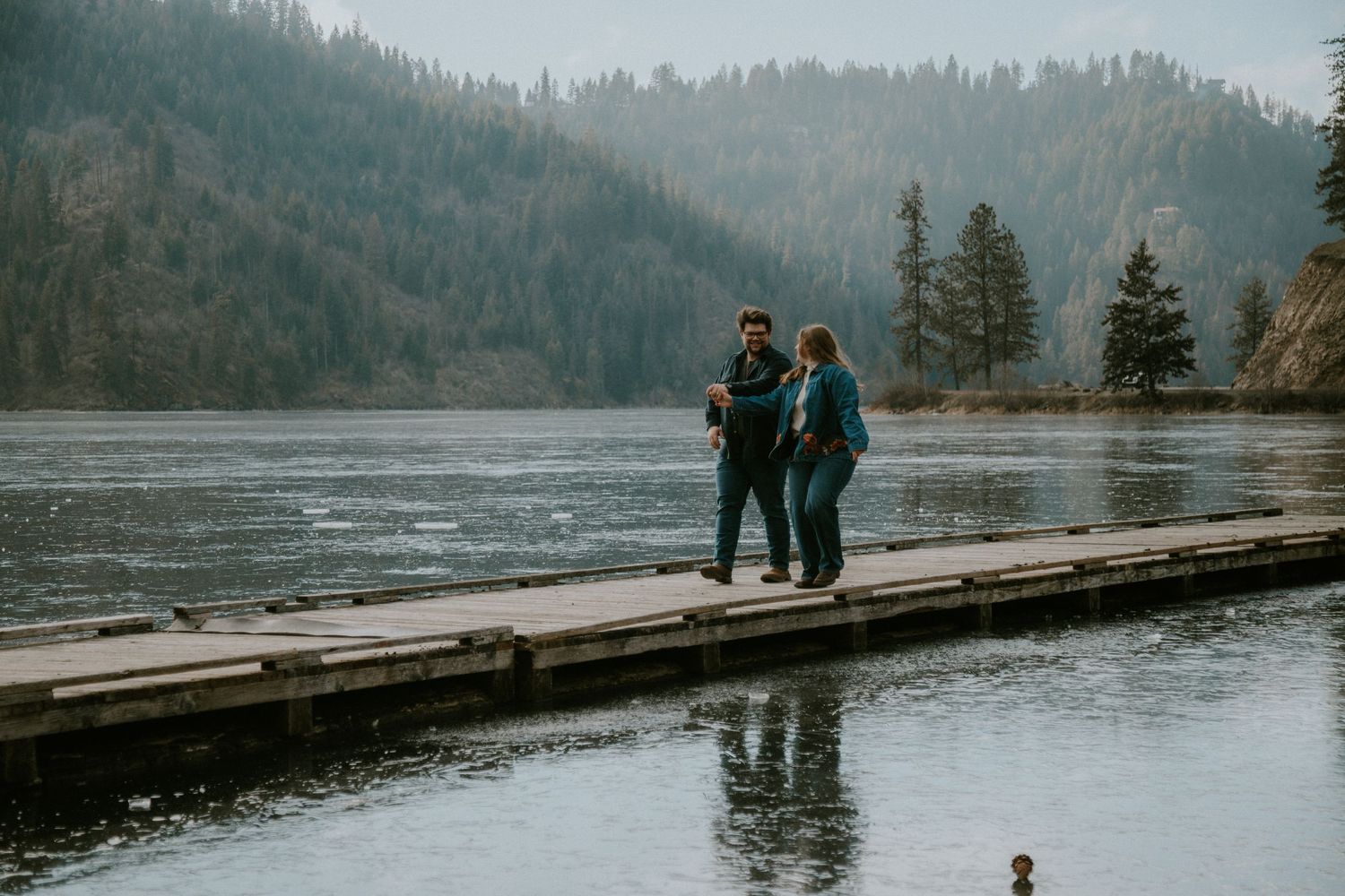 Two people standing on a wooden dock overlooking a river surrounded by pine trees on a misty day.