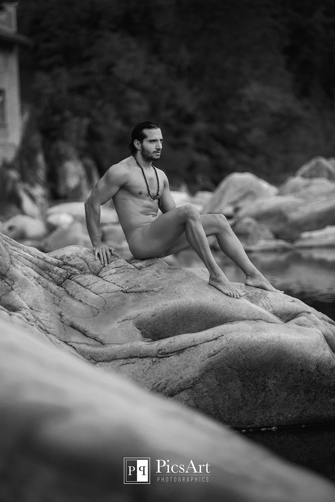 Black and white fine art nude male model posed on smooth rocks in a natural outdoor setting.