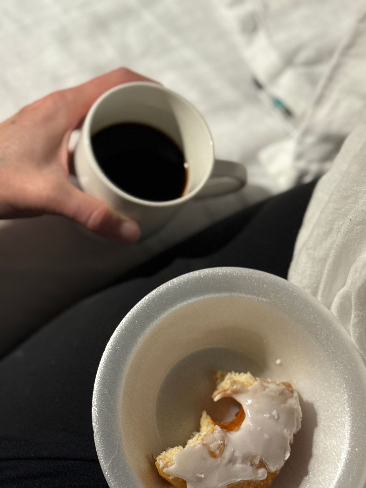 A hand holding a mug of black coffee, with a pastry on a plate in the foreground.
