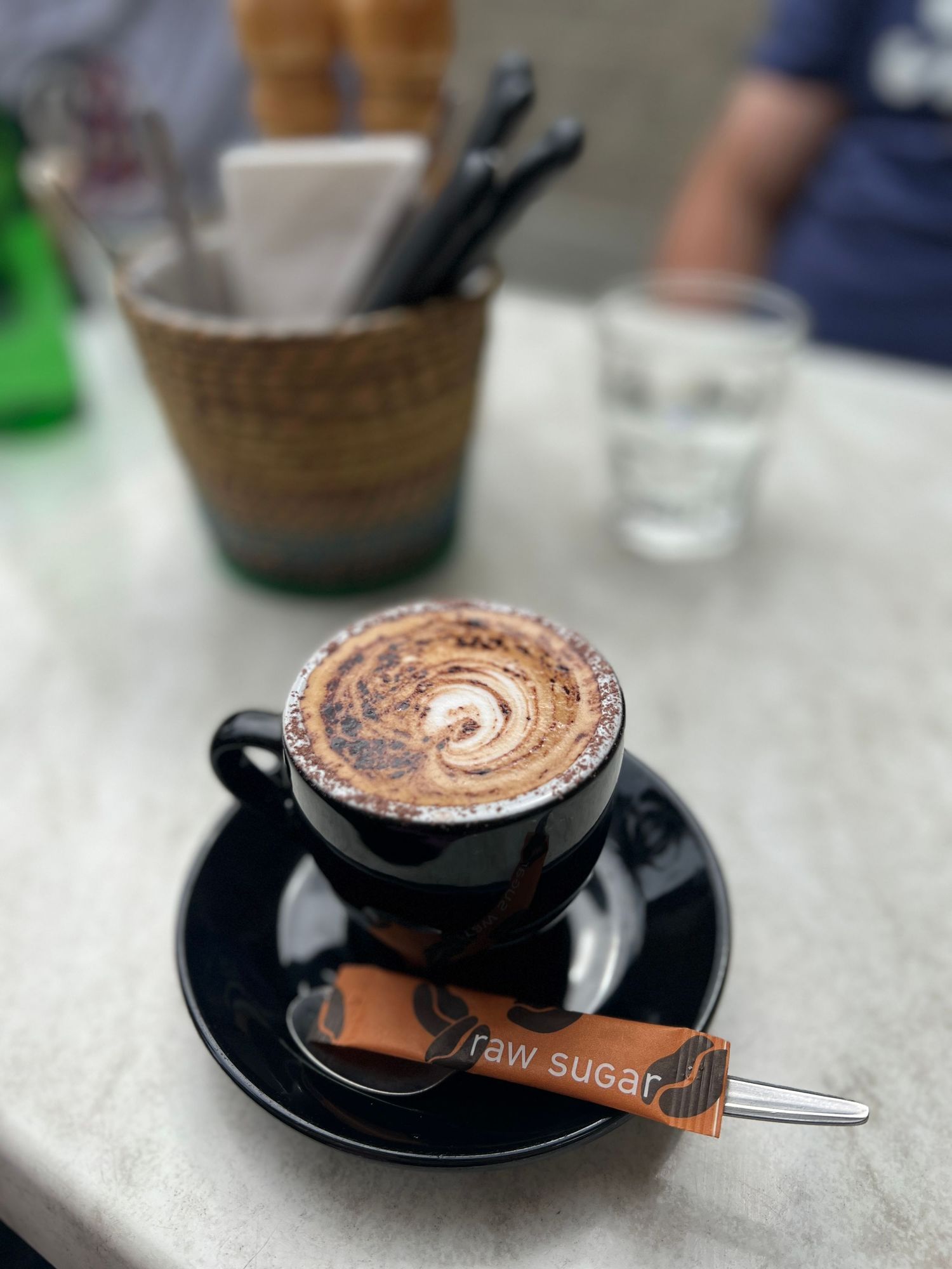 A close-up view of a frothy, swirled cappuccino drink served in a black cup on a saucer with a spoon and raw sugar on a table.