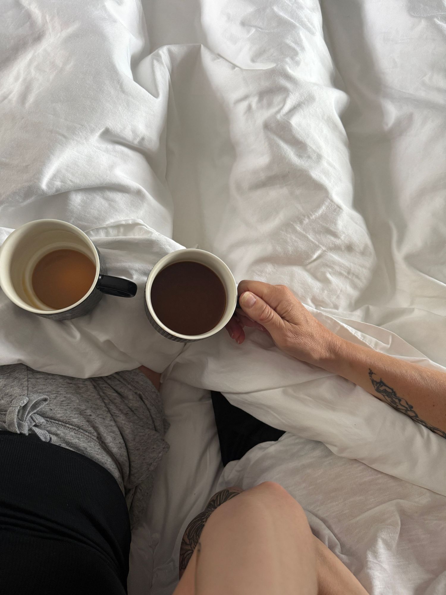 Two hands holding coffee cups on a bed with white sheets.