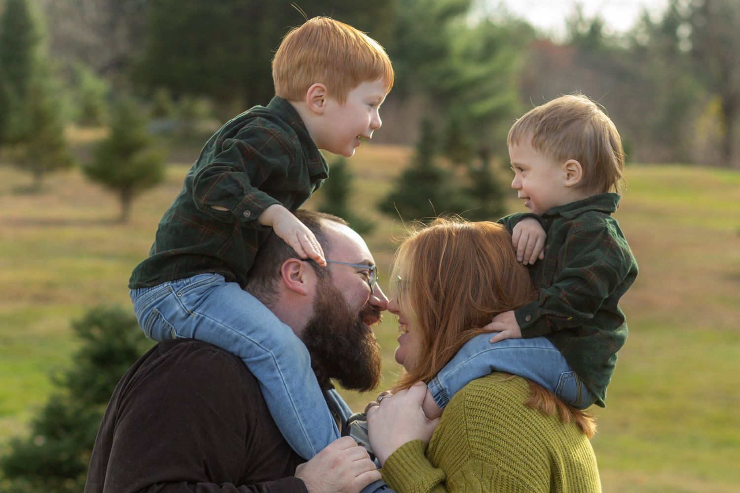 Mother and father with toddlers on their shoulders share a family moment at a Christmas tree farm.