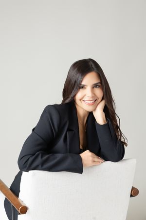 Professional portrait of someone in a black blazer leaning on a white chair against a light grey background.