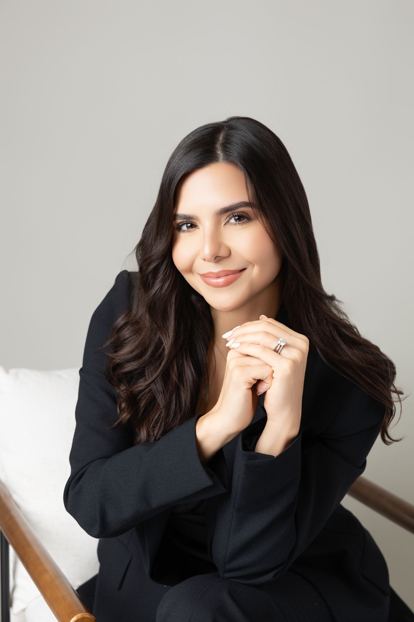 Professional portrait of someone in a black blazer sitting against light gray background with a warm smile.