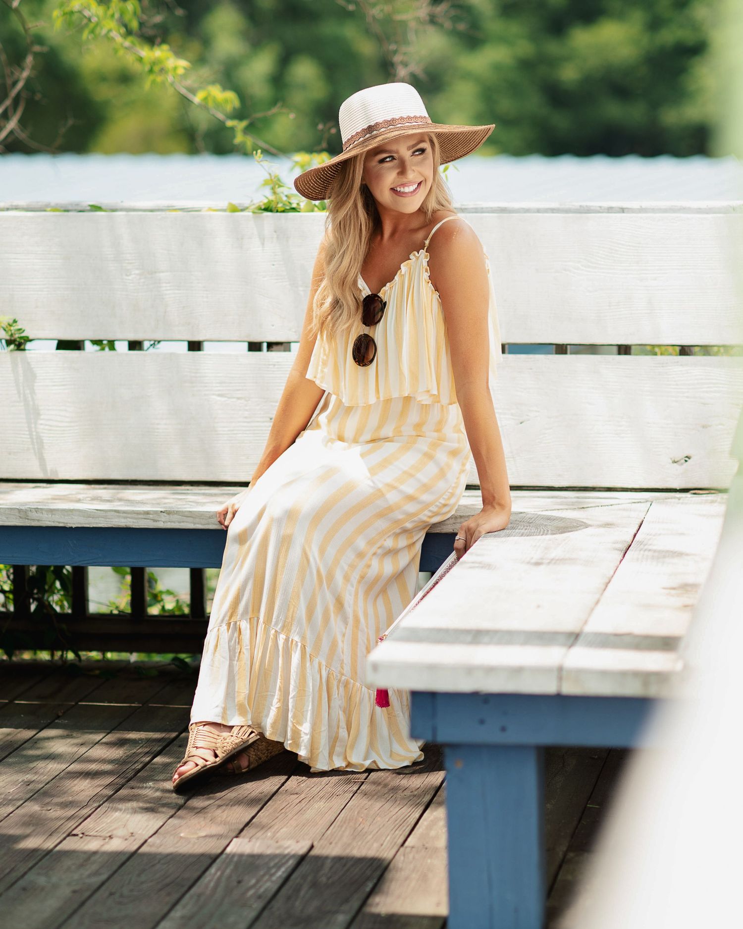 Woman in yellow striped maxi dress and sun hat sits on a blue wooden bench outdoors in summer.