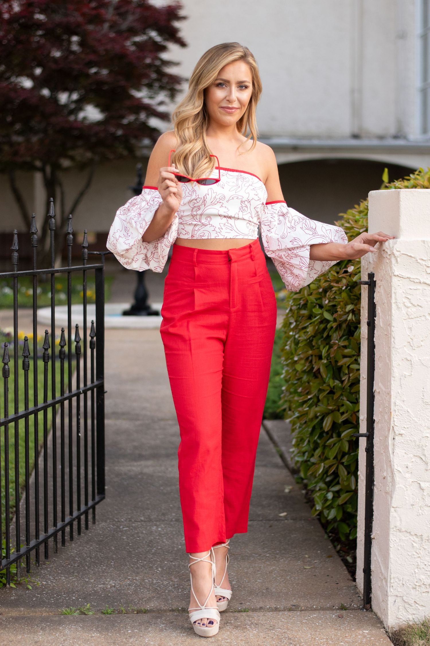 Woman in red high-waist pants and floral off-shoulder crop top with bell sleeves, posing outdoors near iron fence.