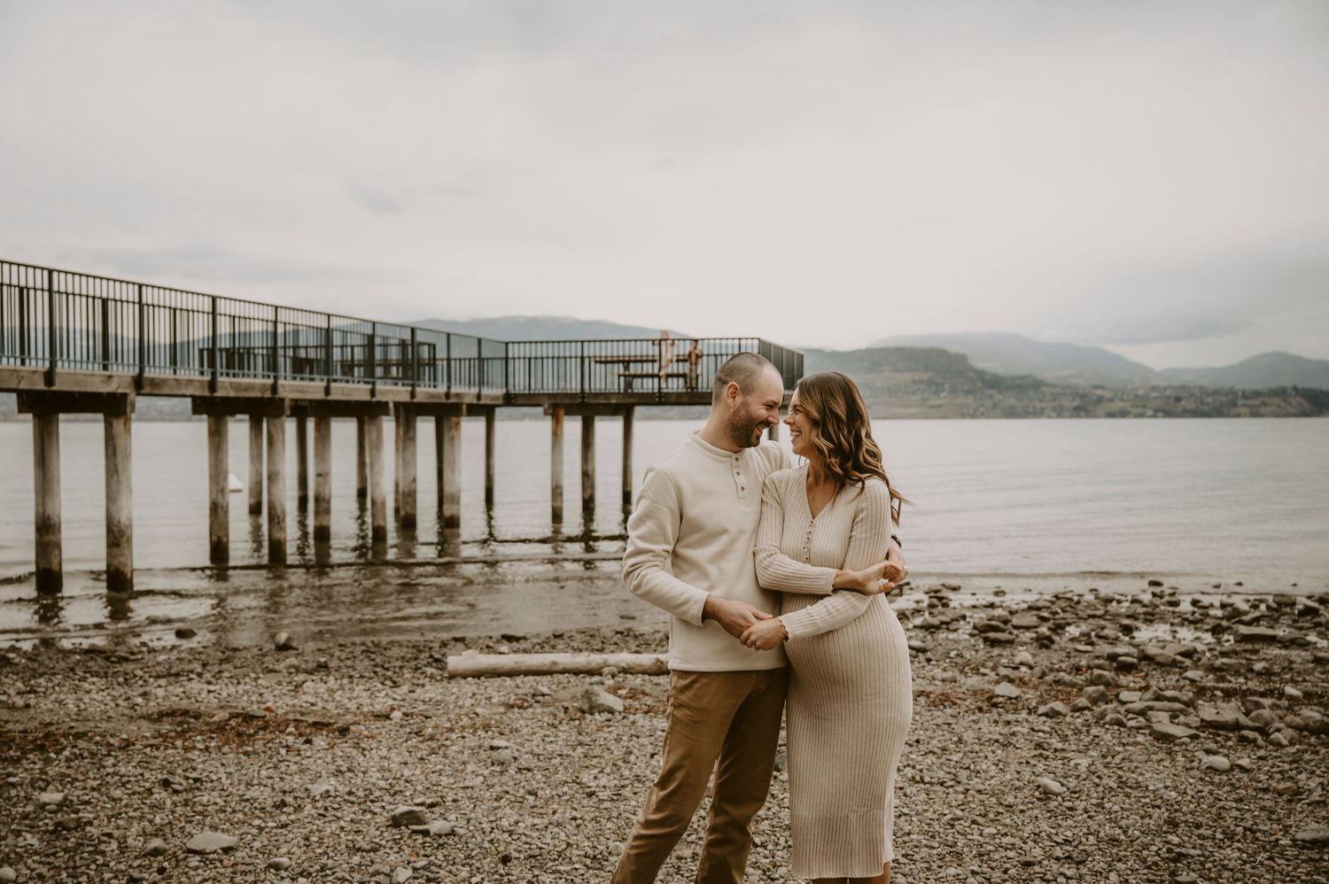 A romantic couple embraces on a rocky beach with mountains and water in the background during sunset.
