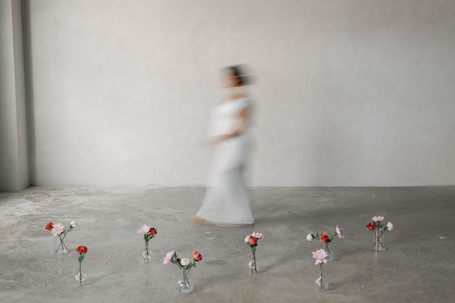 A blurred figure walking in a minimalist, concrete setting with flowers on the floor.