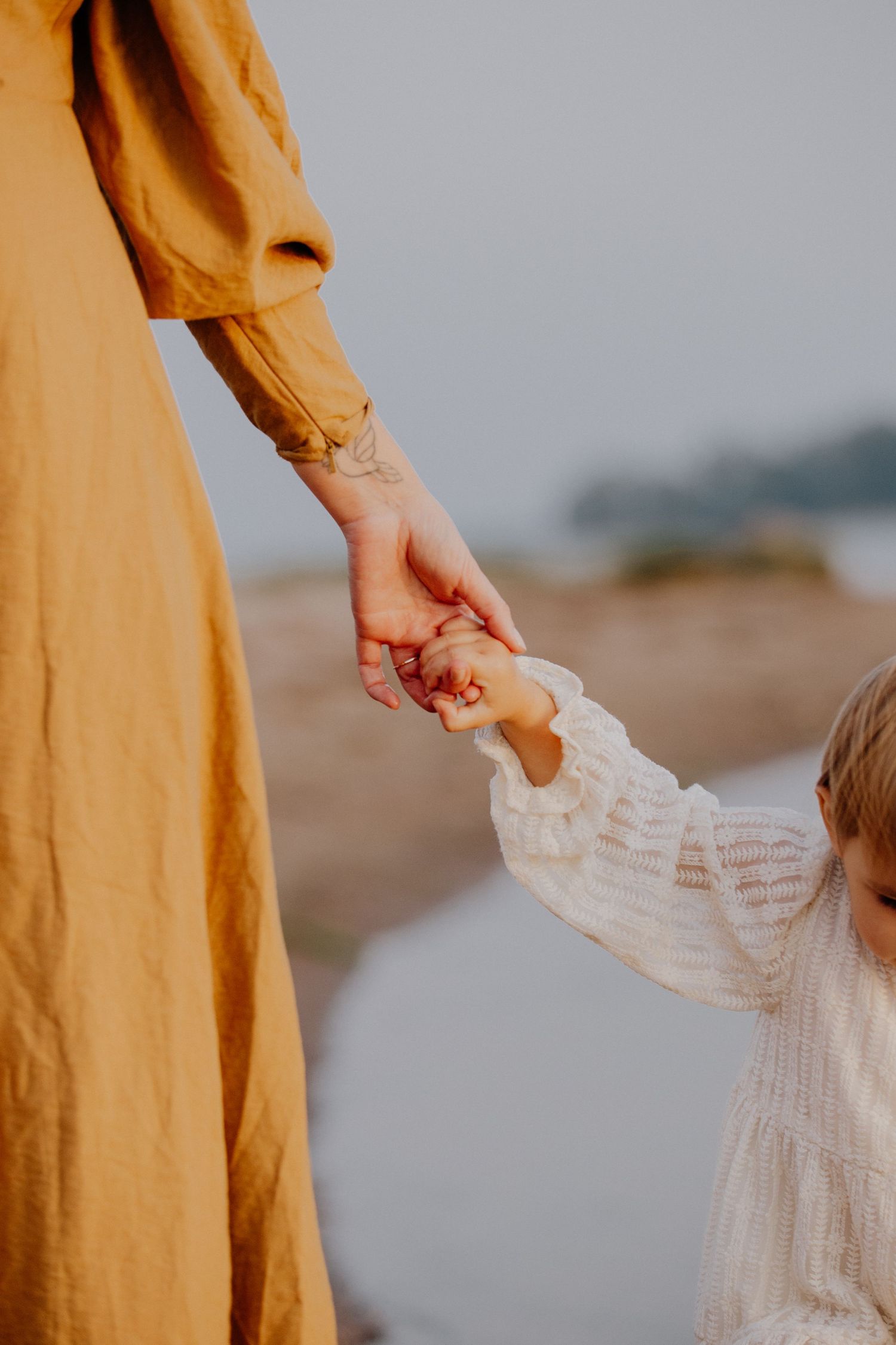 A person in a mustard yellow dress holds hands with a child wearing white on a beach at sunset.