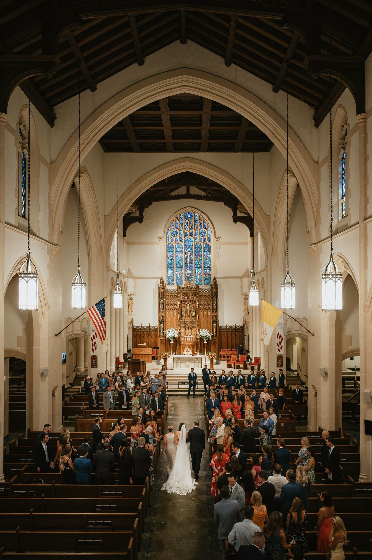 Wide panoramic view of a church wedding ceremony with guests seated in wooden pews under vaulted Gothic arches.
