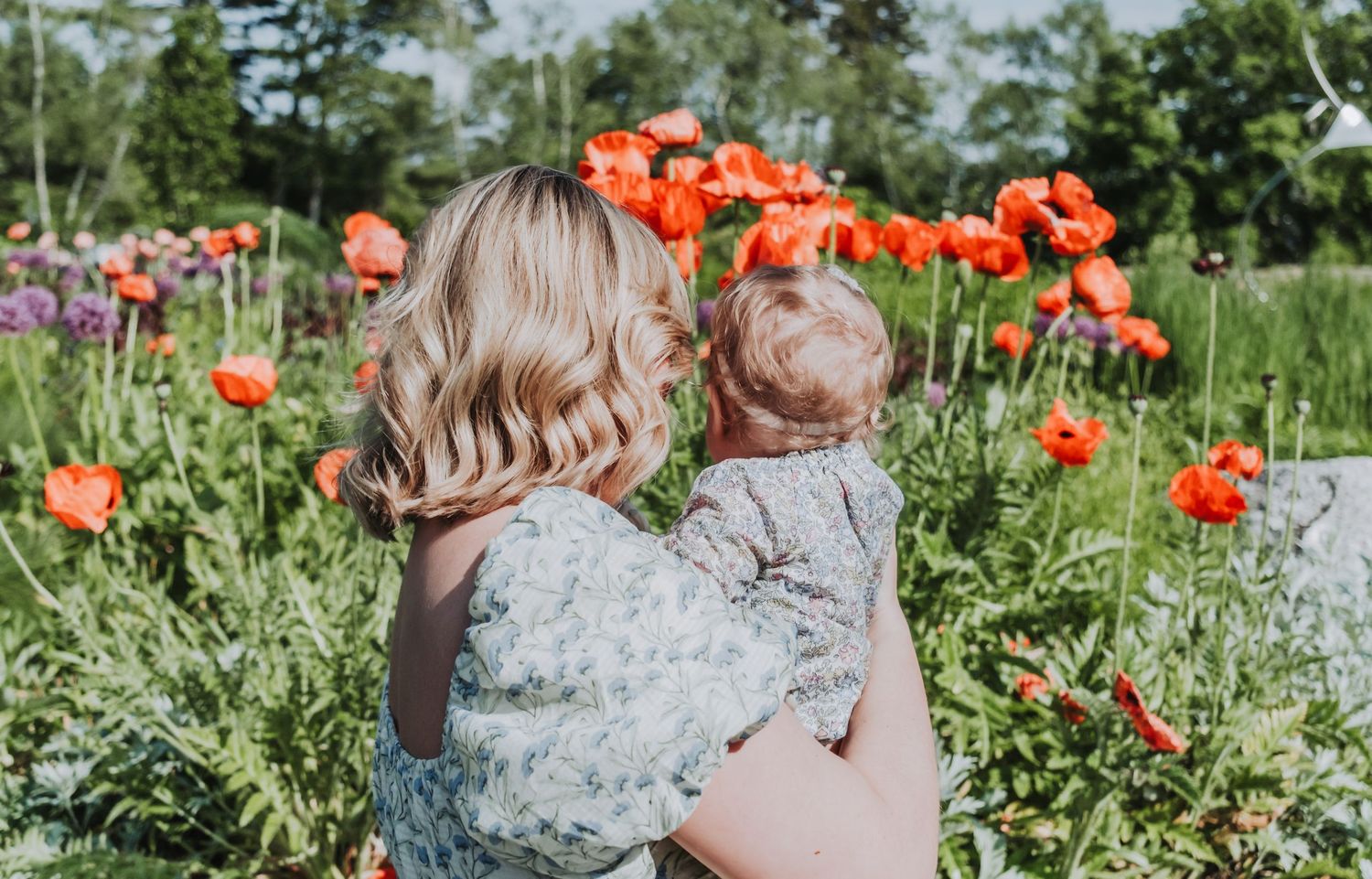 A person in a floral dress stands among vibrant orange poppies in a sunny garden setting.