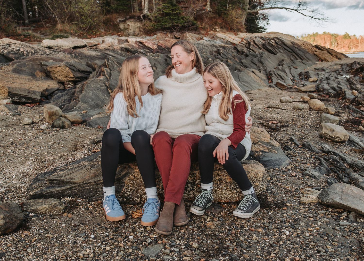 Three friends sitting together on coastal rocks wearing casual sweaters and jeans on an autumn day.