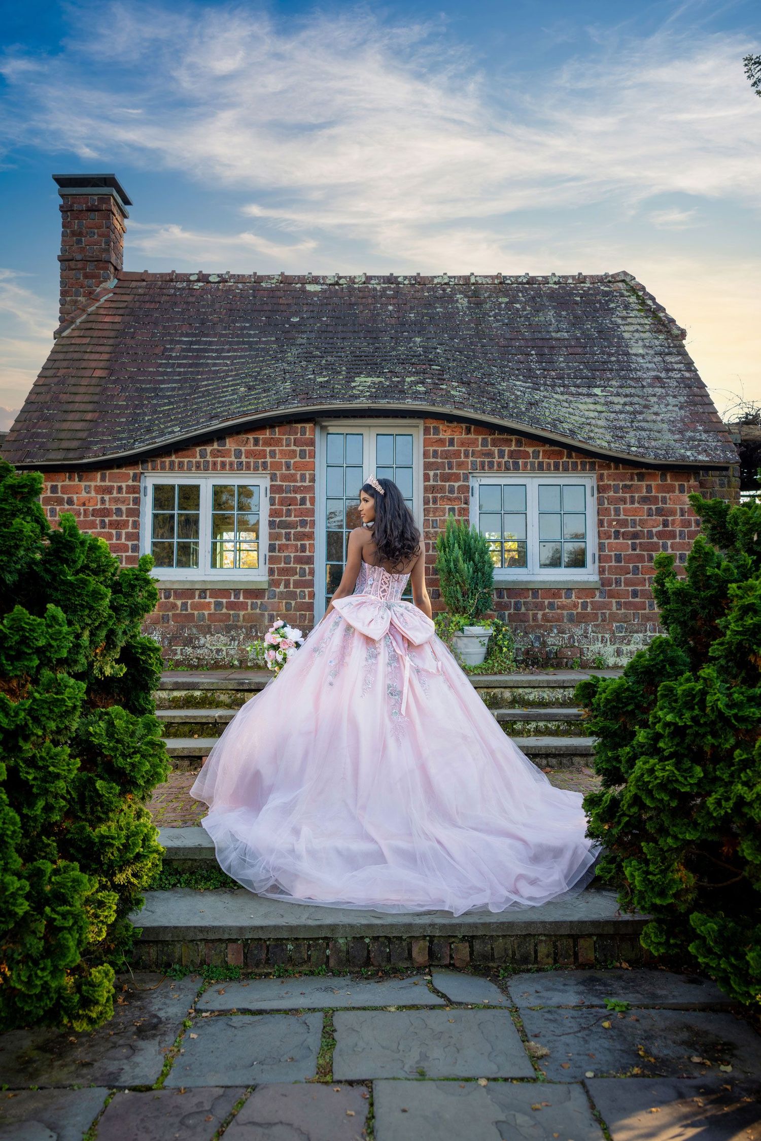 Elegant pink ball gown photographed from behind against a charming brick cottage exterior.