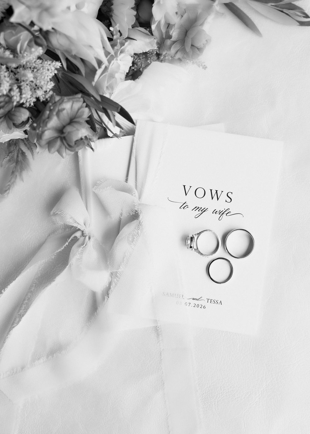 Black and white flat lay of wedding vows booklet, engagement ring, and wedding bands beside a bridal bouquet.