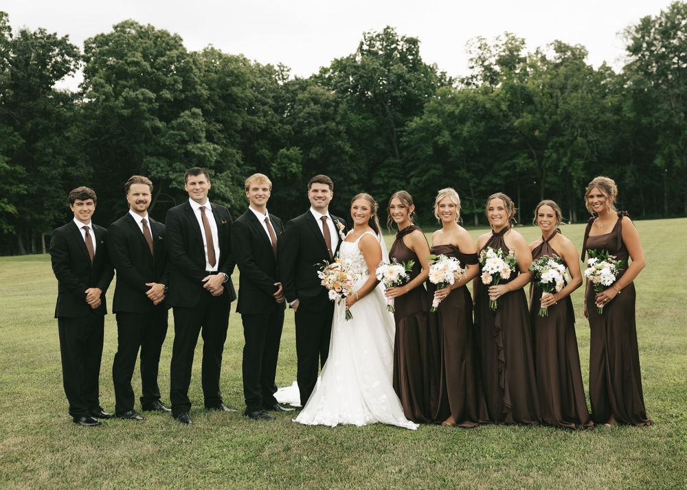 Wedding party poses outdoors on green lawn, groomsmen in black suits and bridesmaids in brown dresses flanking bride and groom.