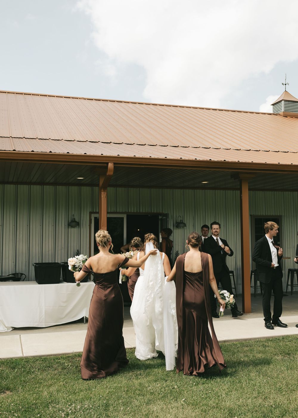 Bride in white gown with bridesmaids in brown dresses outside a barn wedding venue on a cloudy day.