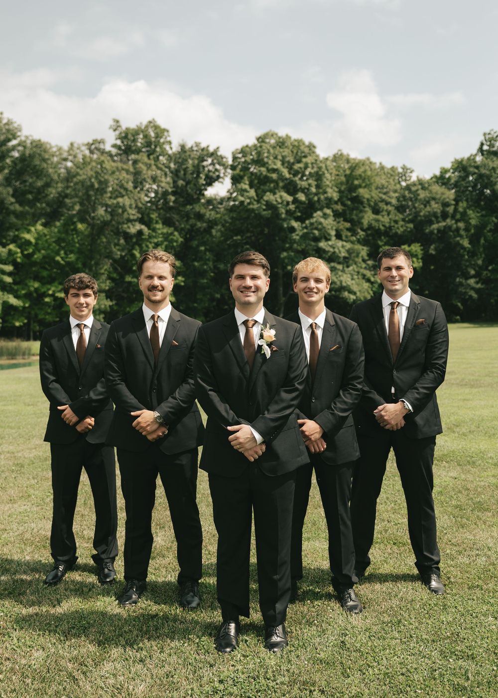 Five groomsmen in black suits with brown ties pose outdoors on green grass with trees in background.