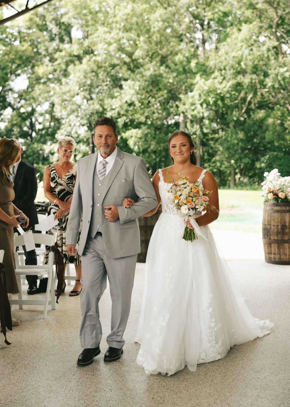 Bride in lace gown walks down aisle with escort holding colorful bouquet at outdoor wedding.