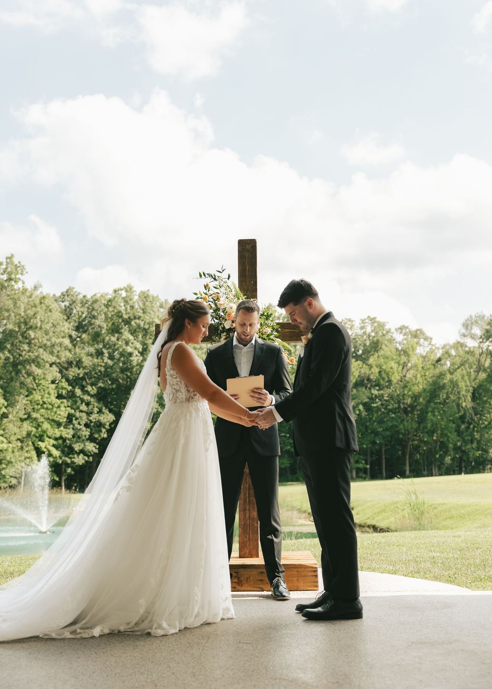 Bride and groom exchanging vows outdoors before a wooden cross with an officiant and lush greenery backdrop.