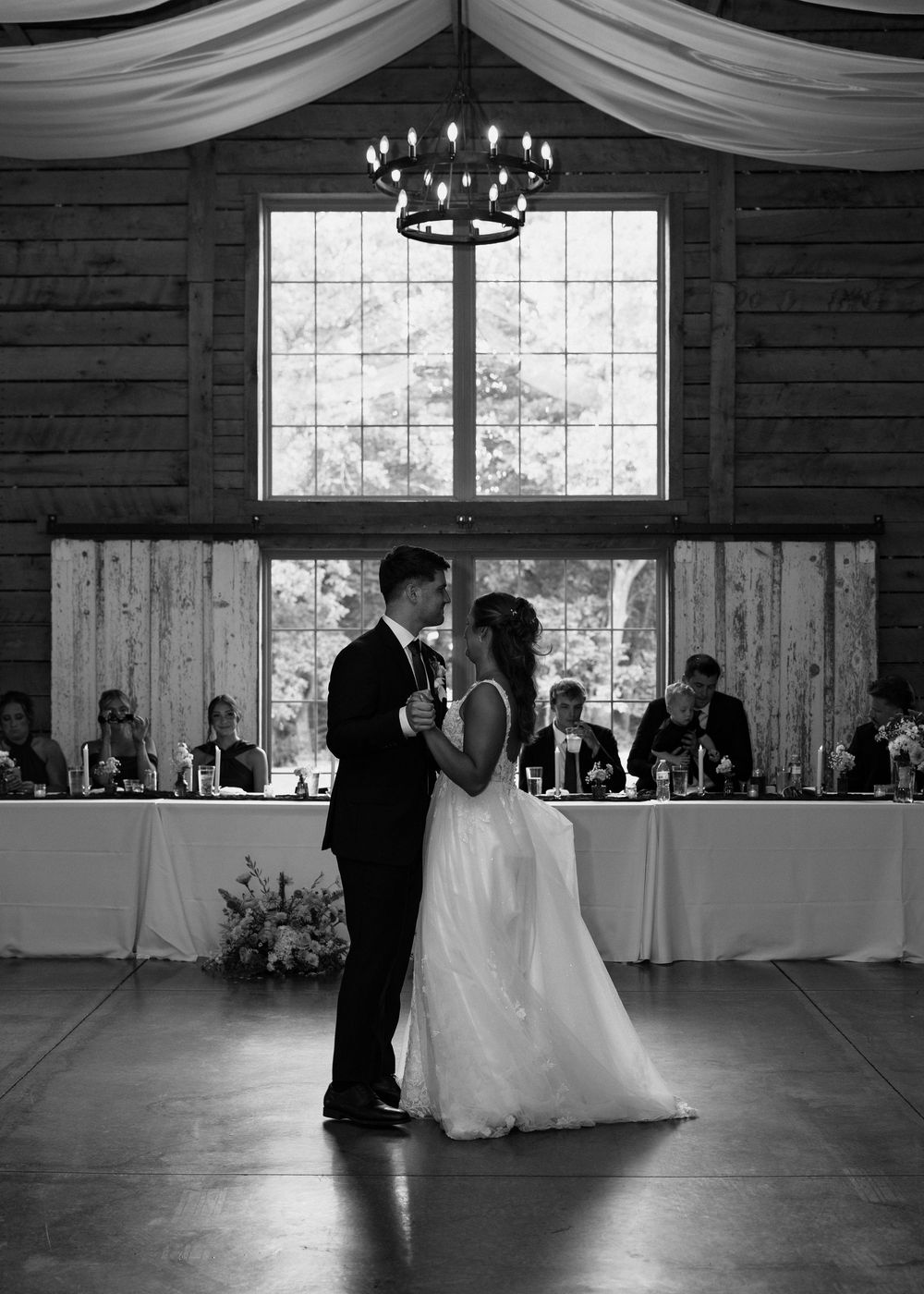 Black and white photo of bride and groom sharing their first dance in a rustic barn venue with chandelier lighting.