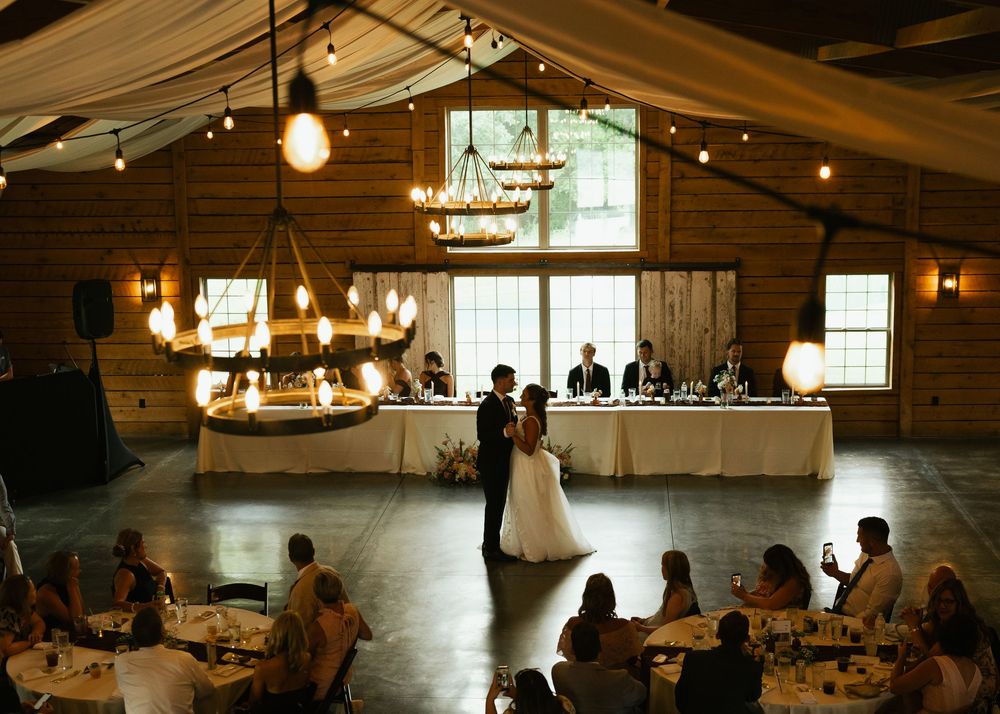 Bride and groom share first dance in rustic barn wedding venue with warm Edison bulb lighting and guests seated at round tables.