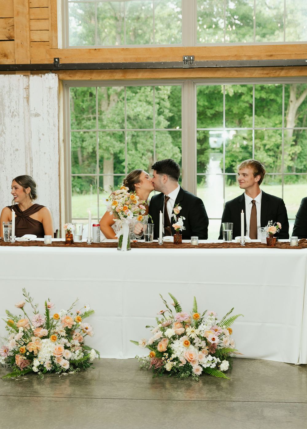 Wedding party at head table with couple kissing, flanked by floral arrangements of pink, white, and orange blooms.